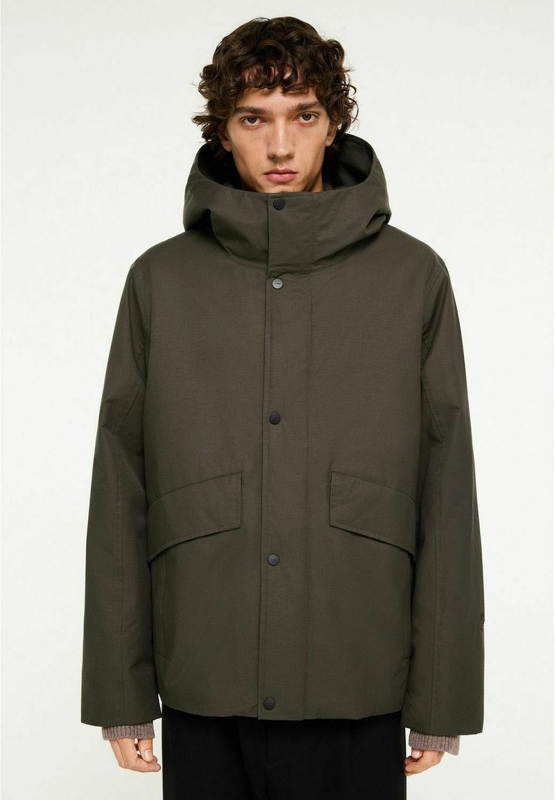 Mörkgrön, huva jacka med lös passform, som har tryckknappar, två framfickor och ribbade manschetter synliga under ärmarna på jackan.