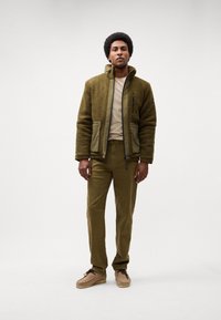 Grüne Fleecejacke mit Reißverschlusstaschen über einem beigen Hemd, kombiniert mit olivfarbenen Hosen und hellbraunen Schuhen. Glatte Textur und lässiges Design.