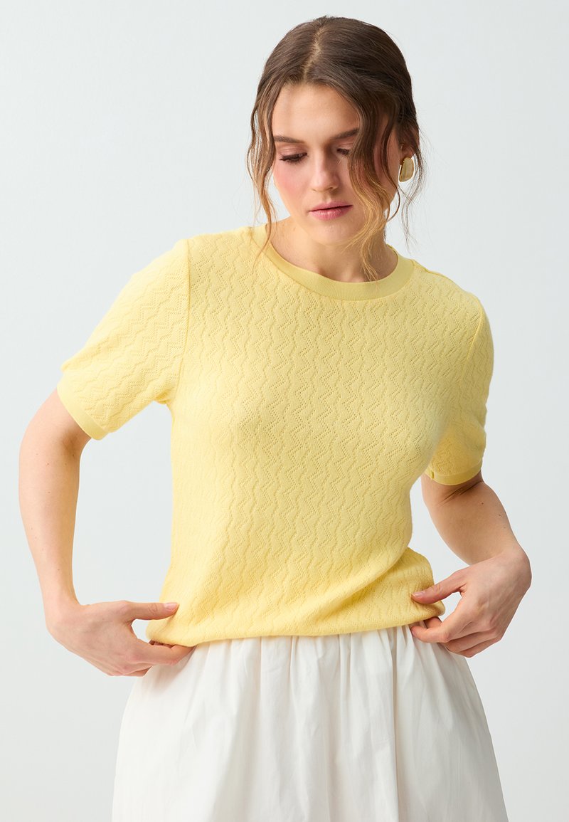 Femme ajustant un haut en maille texturée jaune à manches courtes, portant une jupe blanche, avec les cheveux lâchement attachés et des boucles d’oreilles anneaux dorés sur un fond clair.
