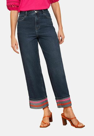 Žena oblečená v tmavomodrých džínsach s rozšírenými nohavicami so sezónnou ružovo-červenou cik-cakovou ozdobou na manžetách a oranžových sandáloch na podpätku, doplnená o jasne ružový top.