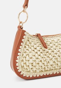 Bolso de paja trenzada con acentos de cuero marrón. Presenta una forma curvada y un anillo de hardware dorado. Cierre de cremallera con un detalle de pestaña lateral.