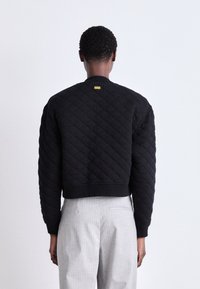 Jaqueta bomber preta acolchoada com colarinho e punhos canelados. Apresenta um rótulo de marca amarelo acima da nuca nas costas. Tecido texturizado.