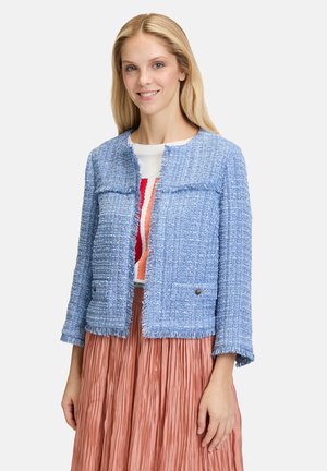 Veste texturée bleu clair avec des poignets et un ourlet à franges, dotée de deux poches avant et de finitions en métal argenté. Portée par-dessus un haut à motifs colorés.