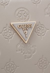 Logo métallique triangulaire Guess USA sur une surface beige texturée avec des motifs floraux et géométriques en relief.