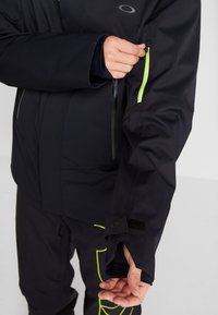 Personne portant une veste noire et un pantalon avec des détails en jaune fluo, utilisant sa main droite pour dézipper la poche de la manche gauche de la veste.