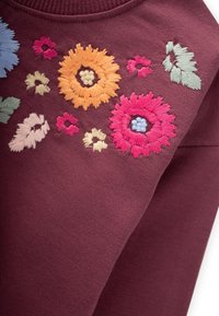 Sudadera burdeos con bordado floral colorido en tonos azules, rosas, naranjas y verdes, que presenta diversas formas de flores y detalles de puntada intrincados.