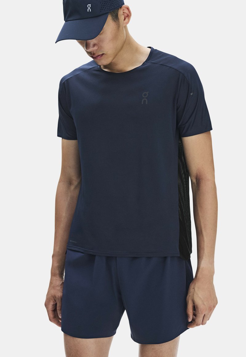 Homme portant une casquette de sport bleu marine, un t-shirt à manches courtes et un short, regardant vers le bas sur un fond clair uni.