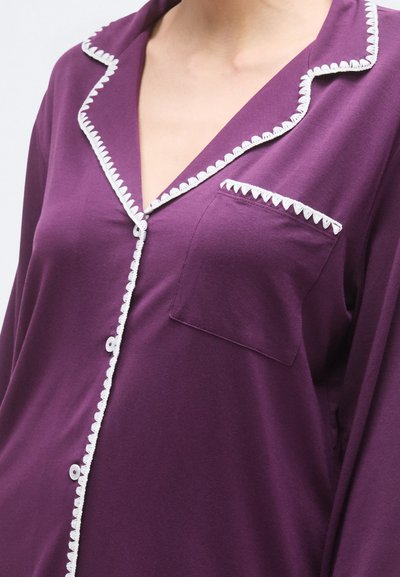 Vijoličasta bombažna pajkama srajca z belim valovitim robom, ovratnikom z zarezom in prsnim žepom. Ima gumbne zapirke spredaj.