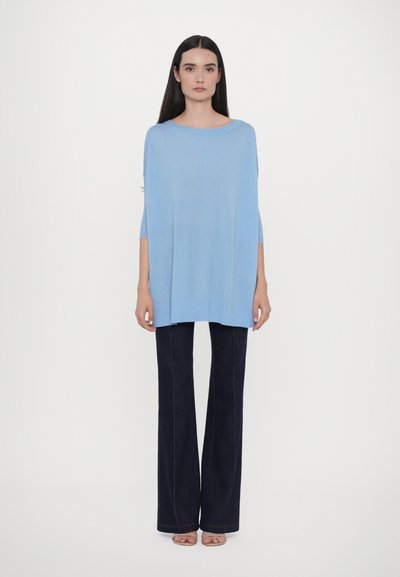 Pulover oversized de culoare albastru deschis cu mâneci până la cot, asortat cu blugi negri evazați, stând pe un fundal alb simplu.