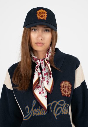 Marineblaues Cap mit goldenem gesticktem Logo, Pullover mit Kragen und beigen Streifen sowie ein floralem Seidenschal mit Nuancen von Burgunderrot und Creme.
