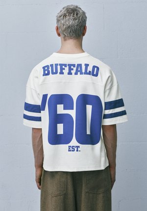 Osoba s krátkými šedými vlasy, otočená zády, v bílém tričku s modrými pruhy a nápisem „BUFFALO '60 EST.“ na zádech.