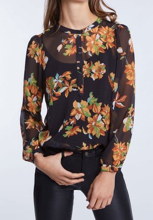 Blouse noire transparente avec imprimé floral orange et vert, col boutonné, manches longues, portée avec un pantalon noir, mains reposant près de la taille.