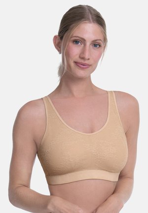 Frau mit blondem Haar in einem beigen Sport-BH mit Spitzenmuster, steht vor einem schlichten hellen Hintergrund und lächelt sanft.