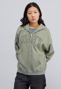 Olive green zip-up hoodie with “OBEY” printed in bold letters. Features a drawstring hood and front pockets. Soft fabric texture.