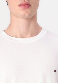 Valge ümmarguse kaelusega t-särk, mis on valmistatud puuvillast, millel on madalal küljel väike punase, valge ja sinise logoga ning sile tekstuur.