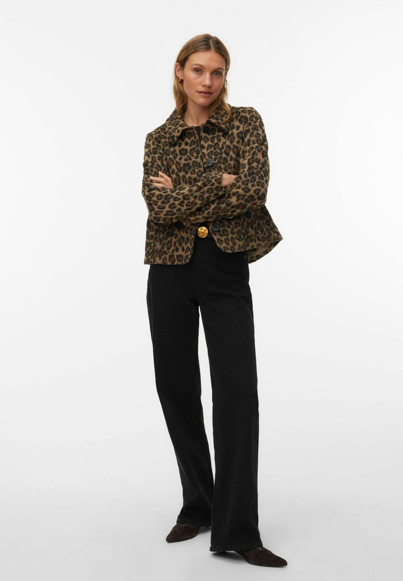 Femme portant une veste imprimée léopard, un pantalon noir avec un bouton doré et des chaussures foncées, debout, les bras croisés, sur un fond blanc.
