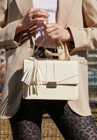 Persona che tiene una borsa in pelle crema con tracolla in oro e ponpon, indossando una giacca beige e pantaloni a stampa leopardo.