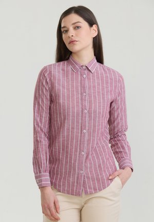 Vrouw draagt een roze blouse met lange mouwen en witte verticale strepen, staat met één hand in de zak van een beige broek tegen een effen achtergrond.