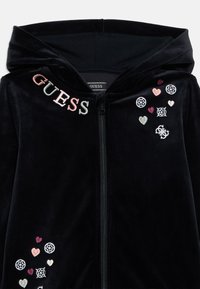 Fekete bársony zipzáros kapucnis pulóver, hímzett "GUESS" felirattal és színes szív- és mintás díszítésekkel az elején.