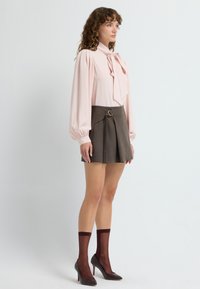 Blusa rosa con un lazo, mangas largas abullonadas, combinada con una falda marrón con plisados que presenta una hebilla circular. Medias de tul burdeos y tacones.