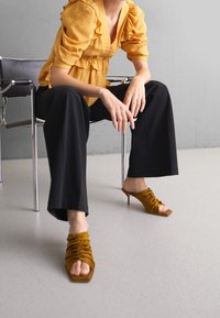 Top giallo con maniche a palloncino e rouches, abbinato a pantaloni neri a gamba larga. Sandali alti con tacco, in marrone, con cinturino e punta quadrata.