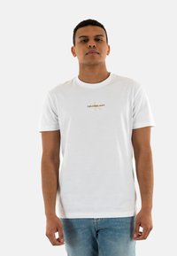 Camiseta blanca de algodón con un logotipo central de Calvin Klein Jeans en dorado, cuello redondo y mangas cortas, combinada con vaqueros azules.