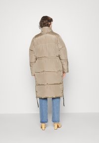 Personne portant un long manteau matelassé beige, un jean bleu et des chaussures à talons beige, photographiée de dos sur un fond blanc.