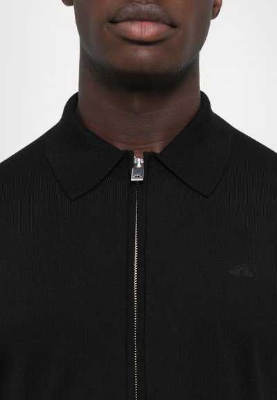 Juodas smulkios struktūros zip-up polo su plokščiu apykakle, turintis sidabrinę užtrauktuką ir subtilų logotipo akcentą krūtinėje. Sklandus audinys ir prigludęs dizainas.