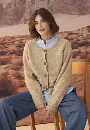 Beige uld cardigan med træknapper, ribbede manchetter og to forlommer, båret over en blå og hvid stribet skjorte, parret med vide denim-bukser.