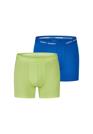Mann richtet leuchtend gelbe Boxershorts, sichtbar vom mittleren Oberkörper bis zur Mitte des Oberschenkels, stehend vor einer Steinmauer mit Pflanzen in der Nähe.