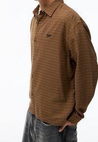 Camisa de manga larga a cuadros marrones, hecha de tela suave, con botones, cuello y un pequeño logo. Combinada con jeans oscuros.