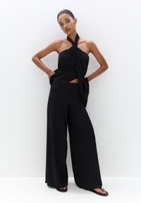 Crni halter top s dijagonalnim drapiranjem i krojenim dizajnom uparen s širokim crnim hlačama. Glatka tekstura, minimalistički stil.