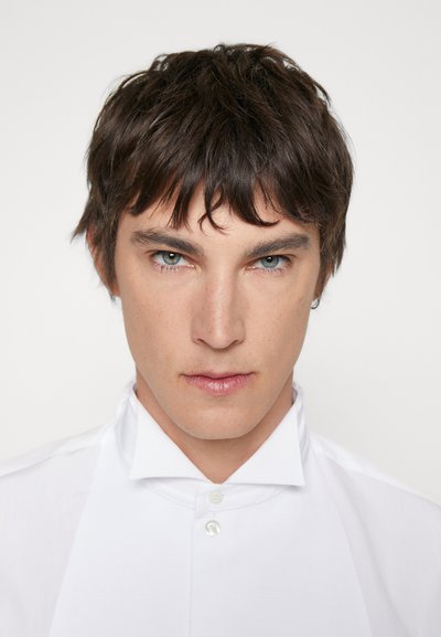 Chemise blanche avec un col haut et un tissu texturé, présentant une fermeture à un bouton au cou, portée par une personne aux cheveux courts et foncés.