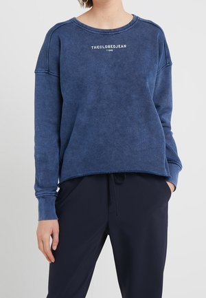 Person trägt ein verblasstes blaues Sweatshirt mit weißem Text "THECLOSEDJEAN 11342" und dunkelblaue Hosen mit Kordelzug an der Taille.