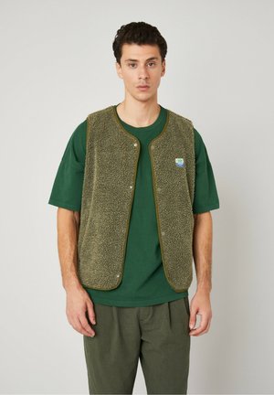 Gilet en polaire vert olive avec une surface texturée, des boutons pression et un biais contrastant. Porté par-dessus un t-shirt assorti et un pantalon décontracté.