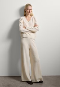 Crème v-hals trui en wijde broek van zachte brei-stof. Relaxte pasvorm met geribde manchetten en tailleband. Donkerbruine schoenen ronden de look af.