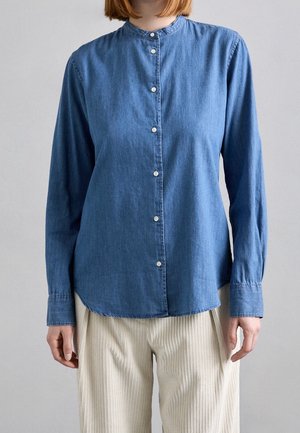 Osoba ubrana w niebieską denimową koszulę z długim rękawem, zapinaną na guziki, z kołnierzem typu mandarynka oraz kremowe spodnie prążkowane.