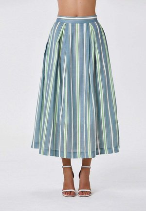 Jupe midi plissée taille haute avec des rayures verticales bleues, vertes et blanches portée avec des talons blancs à bride cheville sur un fond uni.