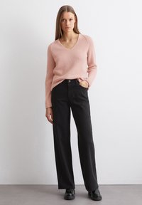 Helles rosa gestricktes Sweatshirt mit V-Ausschnitt, kombiniert mit hochgeschnittenen schwarzen weit geschnittenen Jeans. Schwarze Schuhe runden das Outfit ab.