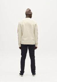 Pulover texturat crem cu manșete cu nervuri, asortat cu pantaloni de culoare albastru închis și adidași. Modelul este văzut din spate, purtând o căciulă maro.