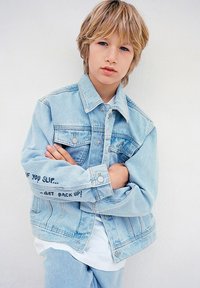 Giacca in denim azzurro chiaro con due tasche frontali e chiusura a bottoni. Presenta una scritta sulla manica: "SE CADI... -RIALZATI!" Texture liscia.
