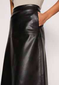 Femme portant une jupe noire en similicuir coupe trapèze avec poche latérale, main placée dans la poche, associée à un haut beige sans manches.