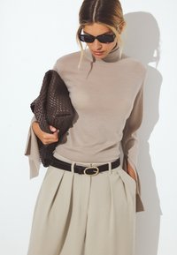 Beige-neulepusero, jossa on kellotettu hihansuu, yhdistettynä vaaleanbeigeihin pleat-takkeihin ja mustaan vyöhön, jossa on kultainen solki. Pitää käsissään ruskeaa ohutta laukkua.