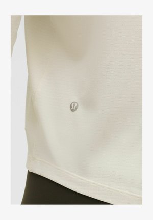 Top atlético de color beige claro, hecho de una tela texturizada, con un pequeño detalle del logo en la parte frontal y un dobladillo curvado.