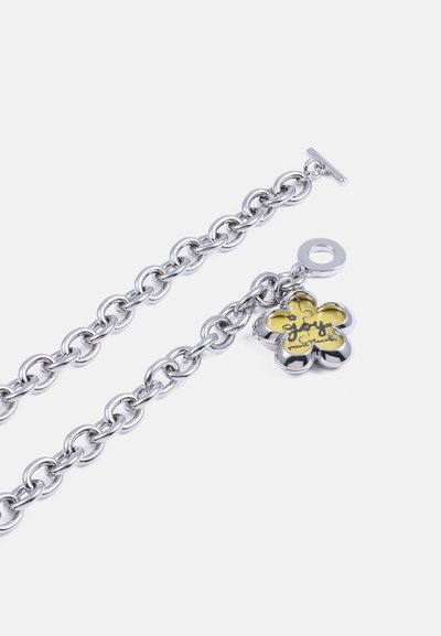 Bracelet en chaîne argentée avec de grosses maillons ovales et un charm en forme de fleur avec de l'émail jaune et un texte en noir à l'intérieur.