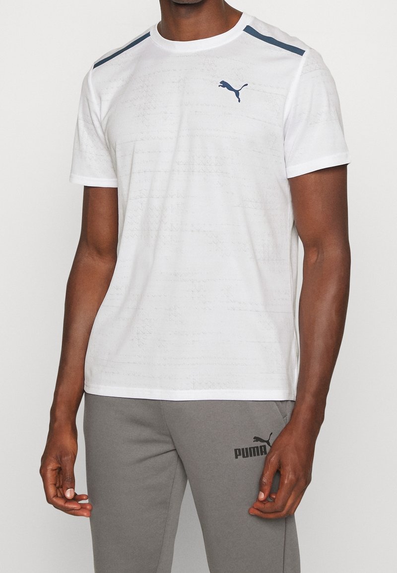 Homme portant un t-shirt de sport blanc Puma avec un logo bleu marine et un pantalon de survêtement gris Puma, debout devant un fond uni.