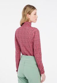Prilagođeni turtleneck top u ružičastom materijalu s uzorkom crnih točkica, uparen s hlačama visokog struka u zelenoj boji. Dizajn na leđima ima duge rukave.