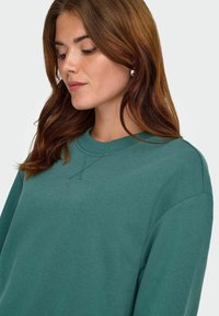 Il sudore color teal presenta un collo a giro, dettagli di cucitura sottili e polsini a coste. Il tessuto ha una consistenza morbida, ideale per un abbigliamento casual.