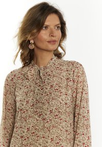 Svetlehnědá blůza s krčním uvazováním, zdobená květinovým vzorem v červené a zelené barvě. Modelka má na sobě kulaté, průhledné náušnice.