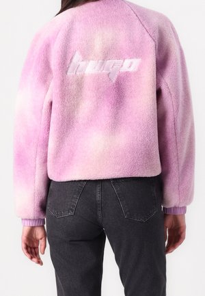 Osoba ma na sobie różowo-białą kurtkę z polaru tie-dye z logo "hugo" na plecach, połączoną z czarnymi jeansami z wysokim stanem.